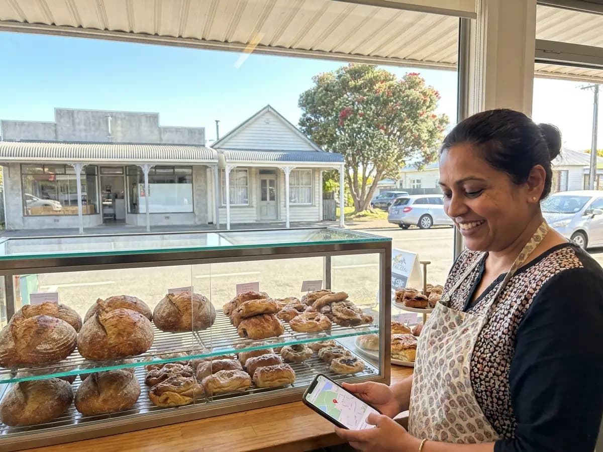 A bakery owner in South Auckland smiling at a map listing on her phone, with a verandah-lined shopping street outside.